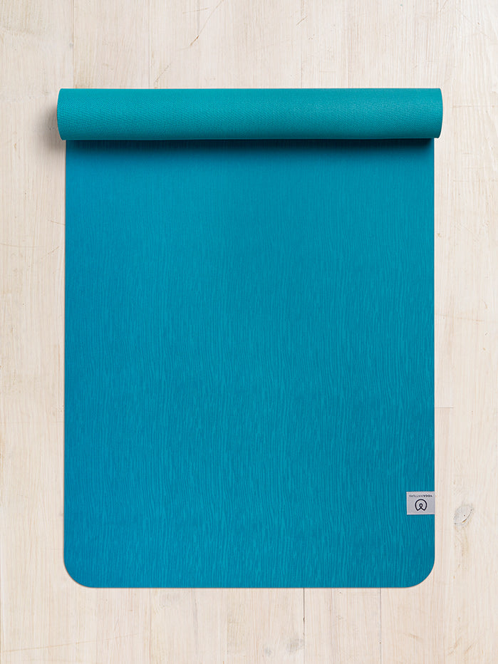 Teal yoga mat with textured surface partially rolled out on wooden floor, top-down view, with visible brand logo.